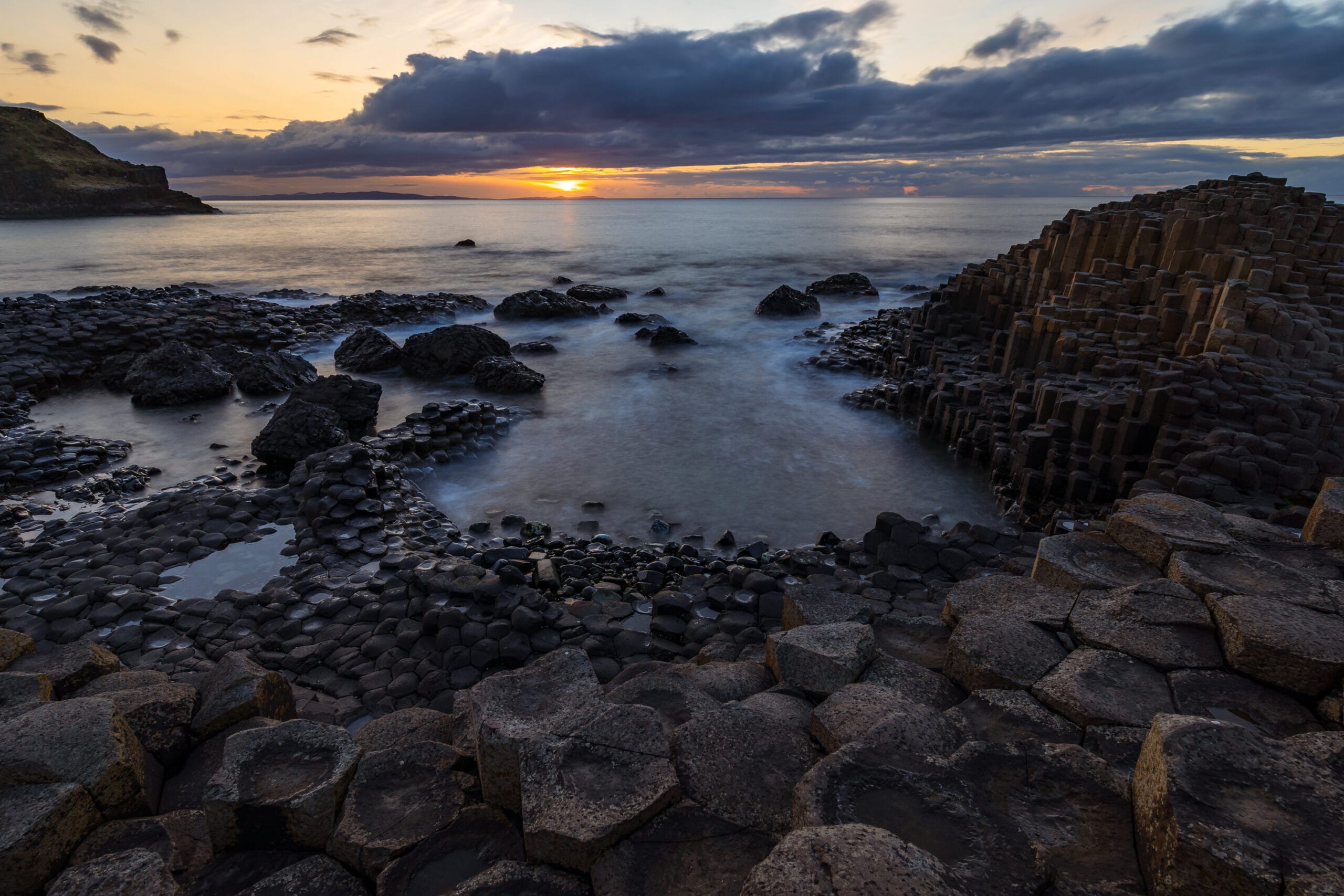 Sunset over the Giant's Causeway in Northern Ireland, with the basalt columns overlooking the sea beneath a dramatic cloudy sky.