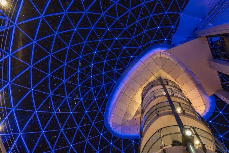 Looking up at the glass dome of Victoria Square in Belfast, showing symmetrical architecture and reflections.