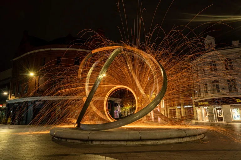 Long exposure steel wool photography in Belfast’s Corn Market, creating trails of sparks around the Spirit of Belfast art piece.