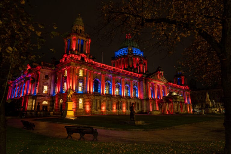 Belfast City Hall illuminated in red and blue at night, framed by autumn trees