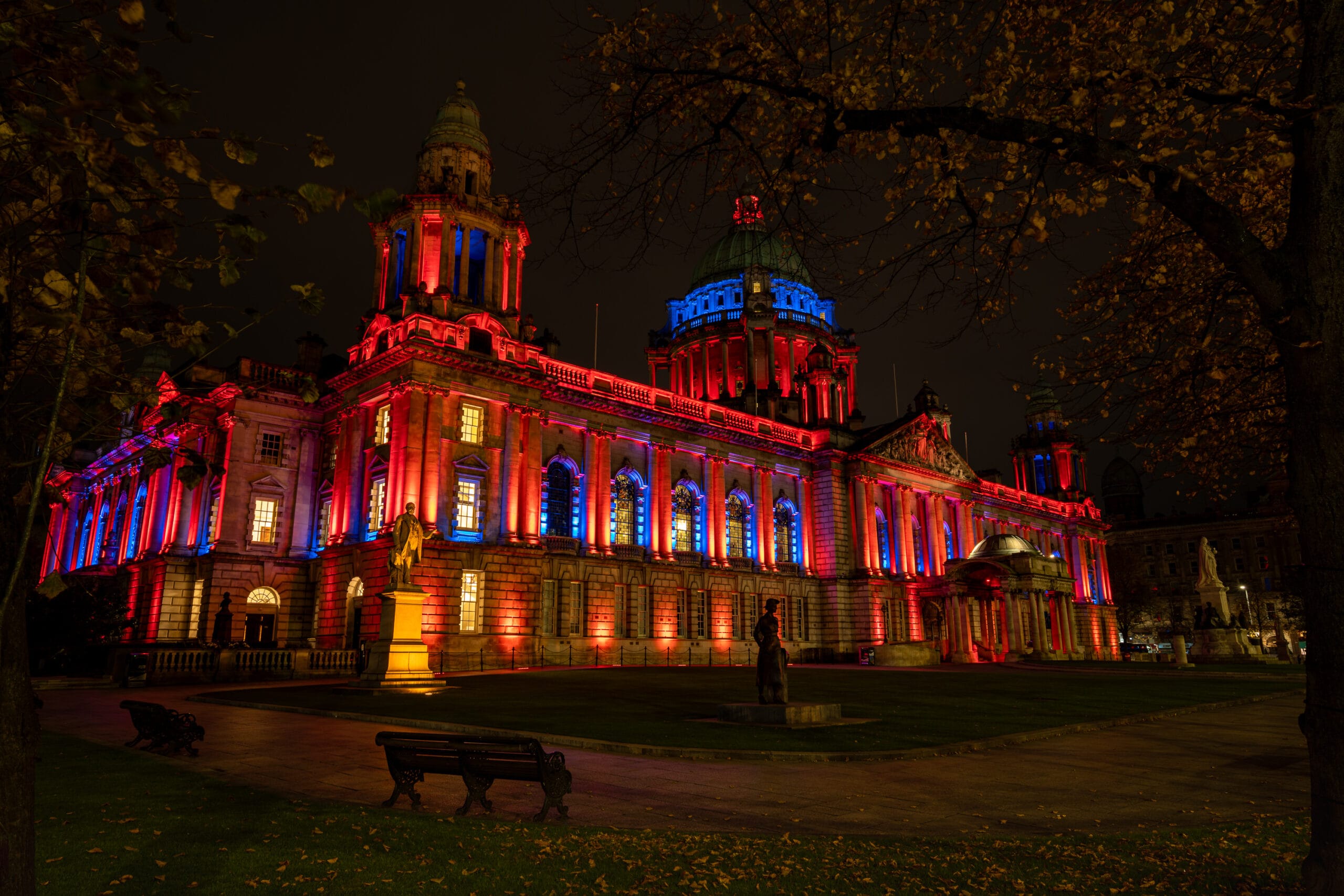 Belfast City Hall illuminated in red and blue at night, framed by autumn trees